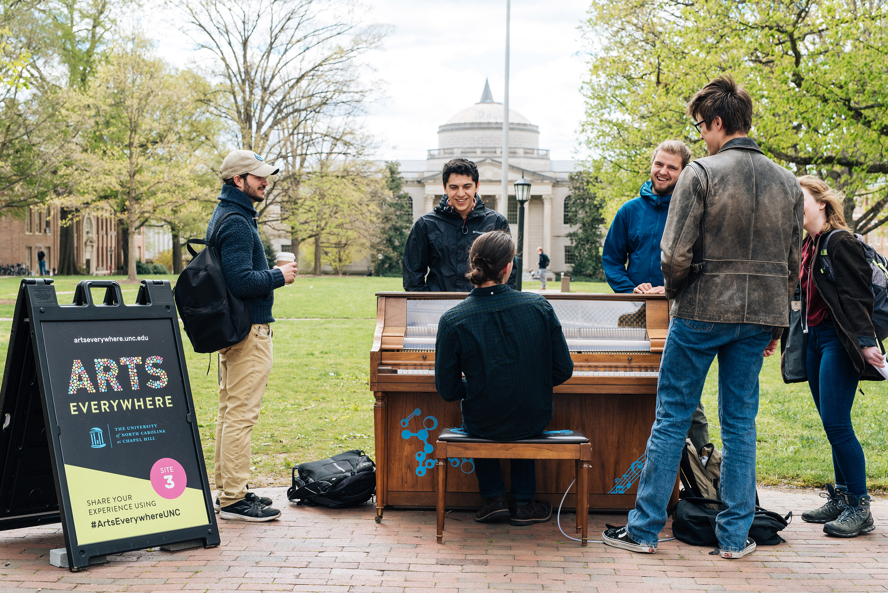 A student is playing a piano on the quad. Five students surround the piano. Wilson Library is in the background.