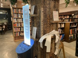 a short story dispenser, which is a black cylinder on the bottom and a glass sheet on top, stands in front of a wooden column. the column is covered in black signatures and has shelves, which hold previously dispensed short stories. bookshelves in the background.