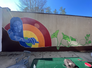 the mural features a woman in blue reaching out to the viewer with a rainbow in the background. Plant life grows around her.
