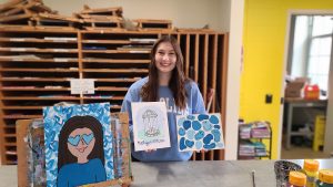 Hannah M. stands in the Morrison Art Studio with three of her painted works